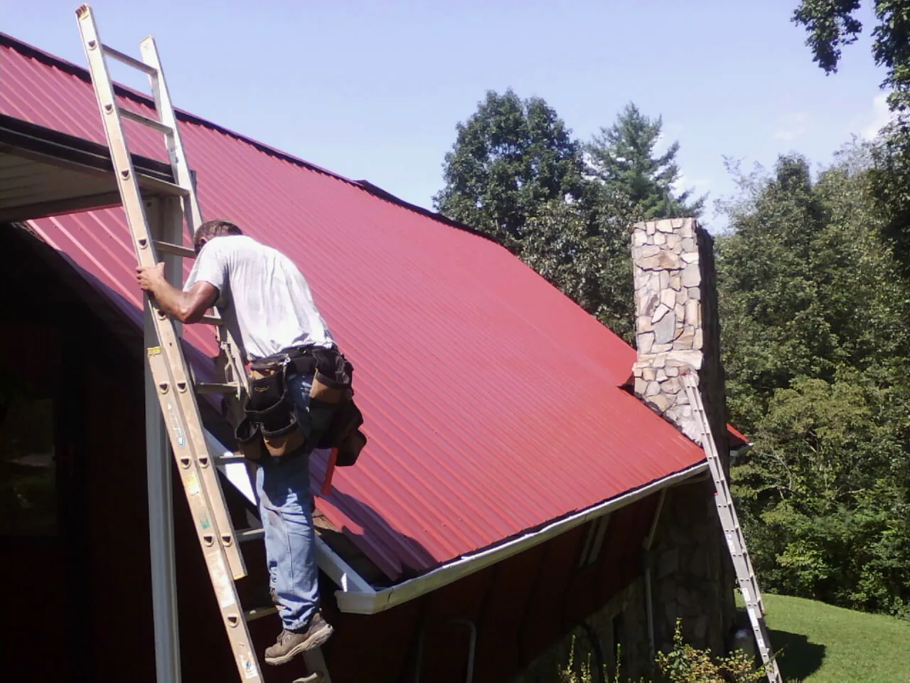 Roofer on a ladder beside a red metal roof and stone chimney — Soffit & Fascia Repair by Roofing Brothers in Phillipsport